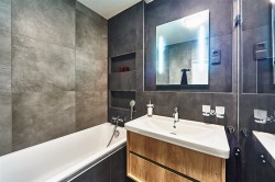 Koupelna s obezděnou vanou, podsvíceným zrcadlem a umyvadlem na dřevěné skříňce; tmavé betonové obklady a nika na kosmetiku ve stěně