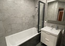 Paneláková koupelna s vanou a černě rámovanou vanovou zástěnou, šedými obklady, bílou umyvadlovou skříňkou a zrcadlovou skříňkou