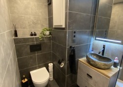 Paneláková toaleta v šedém obkladu: závěsné WC, nika na dekorace a kamenné umyvadlové umyvadlo na dřevěné desce; podsvícené zrcadlo