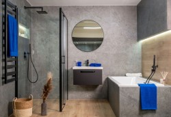 Velká koupelna s betonovým dekorem, vanou obloženou šedými dlaždicemi, závěsnou skříňkou pod umyvadlem a kulatým zrcadlem