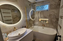 Malá koupelna s oválnou vanou, zlatými profily vanové zástěny, oválným umyvadlem na desce, podsvícenou nikou na kosmetiku a velkým kruhovým zrcadlem