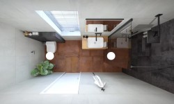 Pohled shora na úzkou koupelnu s umyvadlem na dřevěné desce, sprchou za sklem, terakotovou podlahou a toaletou u okna