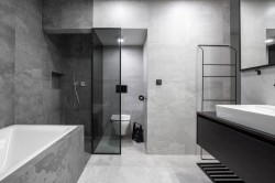 Čistá šedá koupelna s obezděnou vanou, walk-in sprchovým koutem, skleněnou příčkou k WC, tmavou umyvadlovou skříňkou a velkým zrcadlem přes stěnu