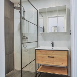 Skleněný sprchový kout s černým rámem vedle umyvadlové skříňky v dřevodekoru; světlé obklady a zrcadlová skříňka nad umyvadlem