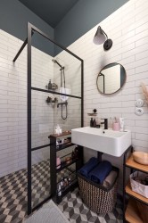 Industriální koupelna s černě rámovaným sprchovým koutem, bílými obklady, kulatým zrcadlem a úložným regálem s kosmetikou u umyvadla