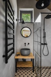 Úzká koupelna se šedou stěrkou, černým žebříkovým radiátorem, umyvadlem na dřevěné otevřené skříňce a sprchovým koutem s černými prvky