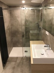 Menší koupelna se sprchovým koutem, umyvadlem na dřevěné skříňce a tmavým žebříkovým radiátorem na šedém obkladu