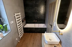 Menší koupelna s vestavnou vanou v tmavém obkladu, podlahou s dřevodekorem, oválným podsvíceným zrcadlem a zlatou baterií