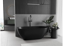 Dominantní matně černá volně stojící vana před šedými obklady, minimalistická černá baterie a přírodní dekorace na kulatém odkládacím stolku