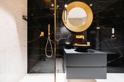 Černá koupelna se zlatými doplňky, kulatým výrazným zrcadlem a umyvadlem na skříňce; walk-in sprchový kout.