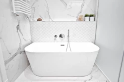 Volně stojící bílá vana v úzkém prostoru, mramorový dekor na stěnách, jemná bílá mozaika za vanou a polička s rostlinami