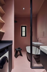 Minimalistická koupelna v pudrově růžové s černou volně stojící vanou, skleněnou zástěnou, policemi nas pračkou a malým oknem