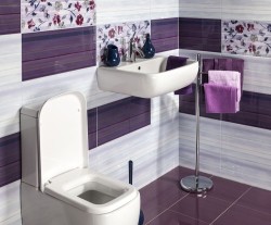 Závěsné bílé umyvadlo a WC v koupelně s obklady v bílé a fialové, doplněné květinovým dekorem a fialovými ručníky