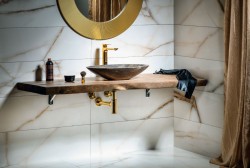 Detail kamenného umyvadla na desce; zlatý sifon i baterie a kulaté zrcadlo ve zlatém rámu na mramorovém obkladu