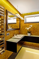 Koupelna se zlatou stěrkou na stěnách, černou rohovou skříňkou, bílým umyvadlem a velkým zrcadlem v dekorativním rámu