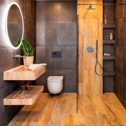 Menší koupelna s tmavými obklady, podsvíceným kulatým zrcadlem, závěsným WC a sprchou s výrazným dřevěným panelem a vestavnými policemi