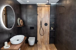 Tmavá koupelna s velkými obklady, oválným umyvadlem na dřevěné desce, černou sprchovou baterií a sprchovým koutem