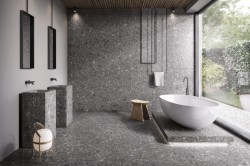 Minimalistický prostor s šedým kamenným dekorem, dvojicí hranatých umyvadel a volně stojící vanou u velkého okna