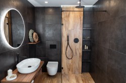 Tmavá koupelna s oválným podsvíceným zrcadlem, bílým umyvadlem na dřevěné desce a sprchou v dřevodekoru s černými prvky