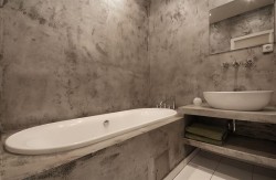 Malá koupelna s vestavnou vanou, otevřenou policí pod umyvadlem a stěnami v surovém betonovém vzhledu