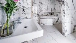 Koupelna s bílým mramorovým obkladem, hranatým umyvadlem se dvěma bateriemi a rohovou vanou