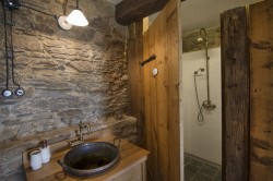 Kamenná stěna a masivní dřevěné dveře u sprchového koutu, kulaté umyvadlo na dřevěné skříňce a mosazné baterie ve venkovském stylu